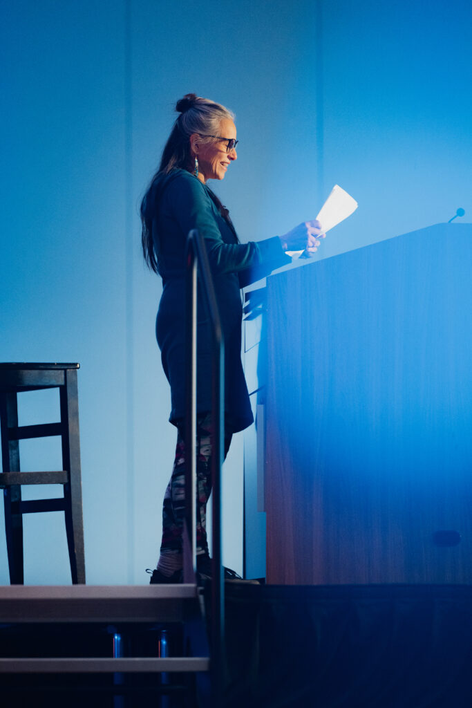 An American Indigenous woman with long, silvery hair in a ponytail, stands in front of a lectern giving a talk to an audience.