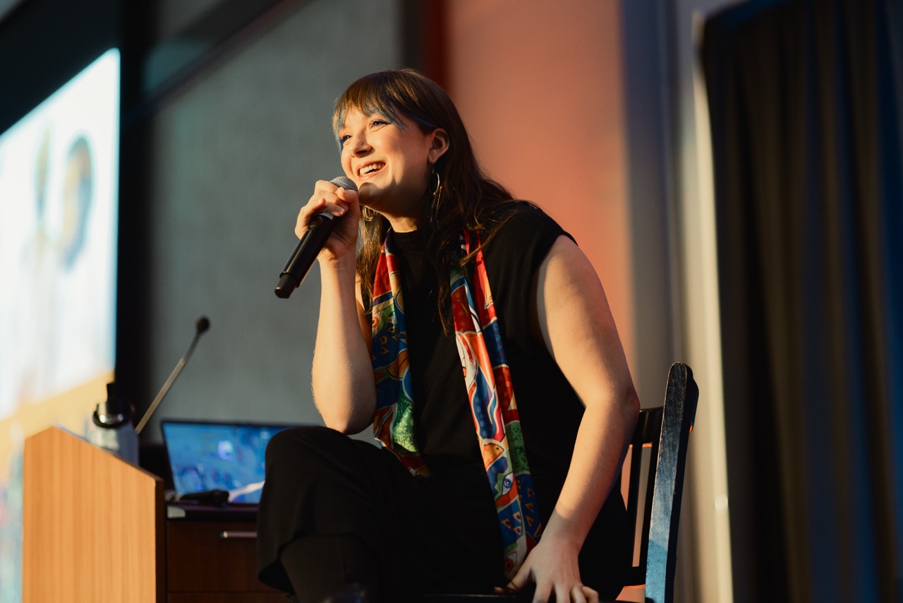 A white woman with vibrant-colored hair smiles while holding a microphone on stage.