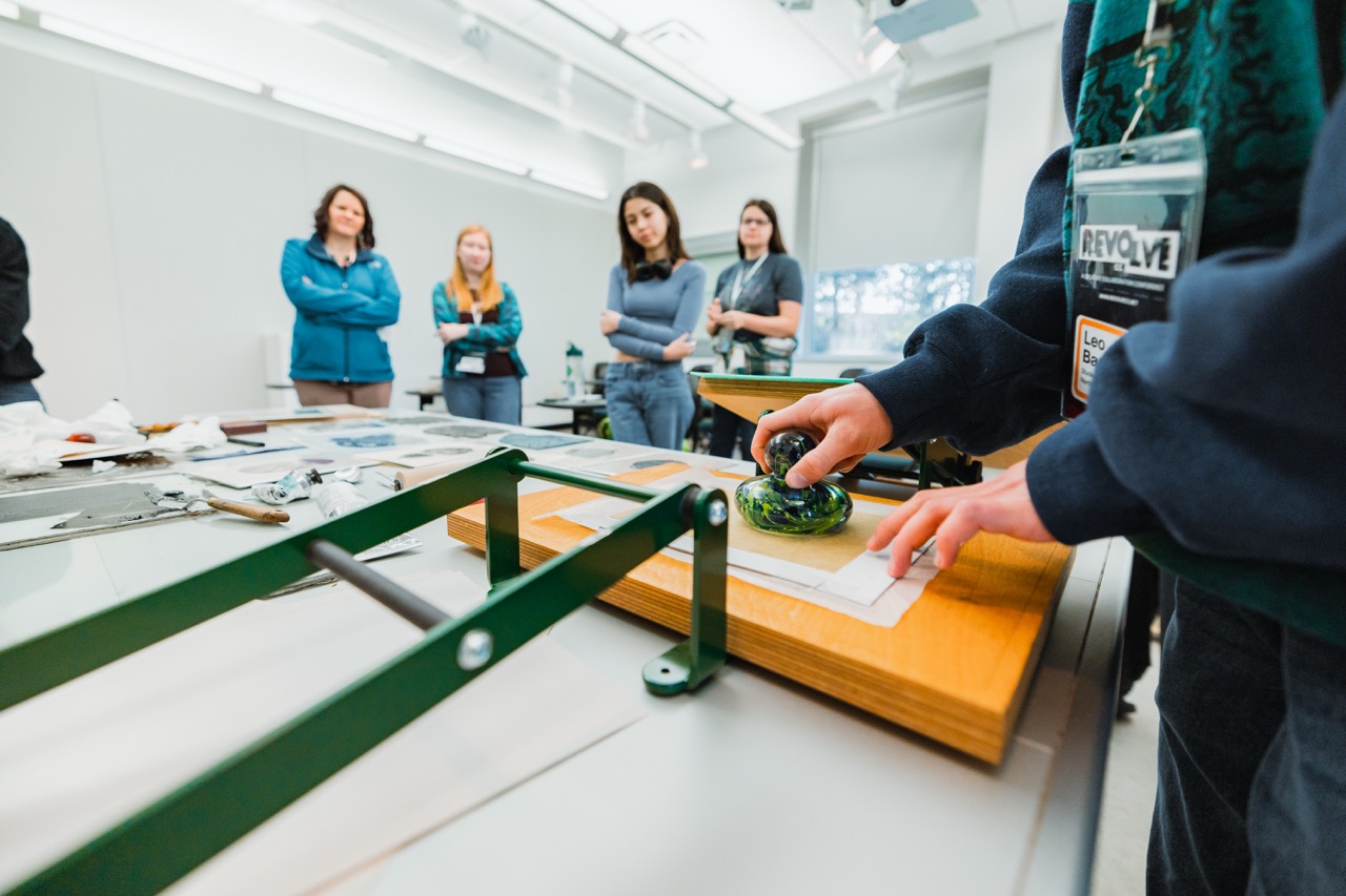 A hand presses down on an ink press, with a small crowd of onlookers standing around.